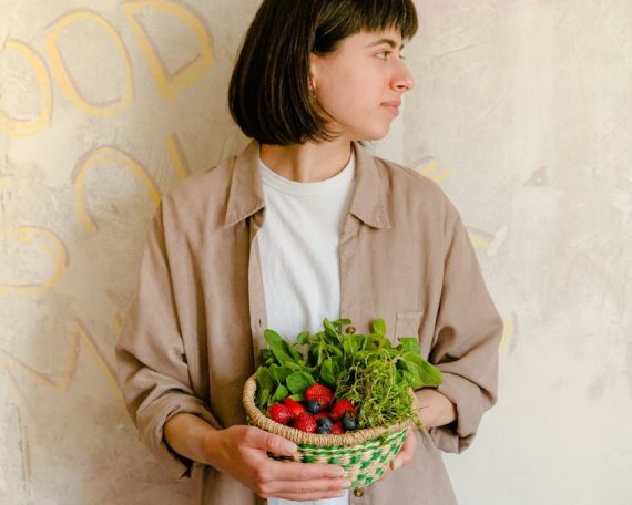 Woman standing with a basket of fresh greens and berries against a rustic wall background.